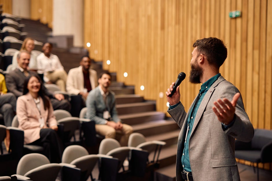 Young Caucasian professor with brown hair and a beard wearing a teal button-up shirt and grey suit jacket speaking to a group of faculty members in an auditorium 
