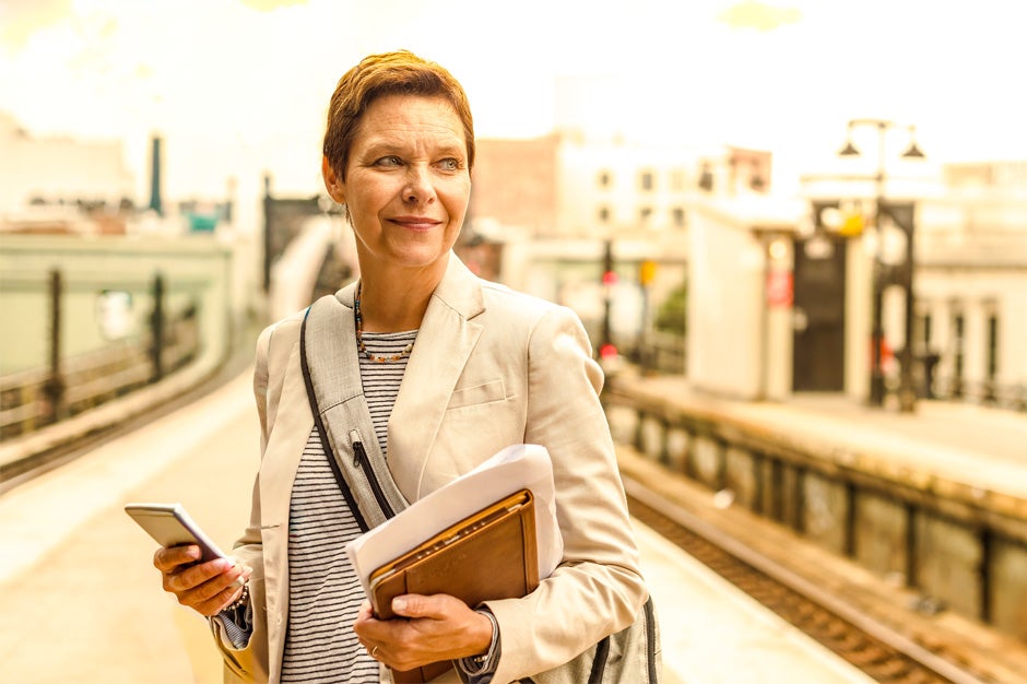 Senior female professor with short red hair, a white and black stripped shirt and cream-colored blazer holding her phone and notebooks outside of a foreign train station 