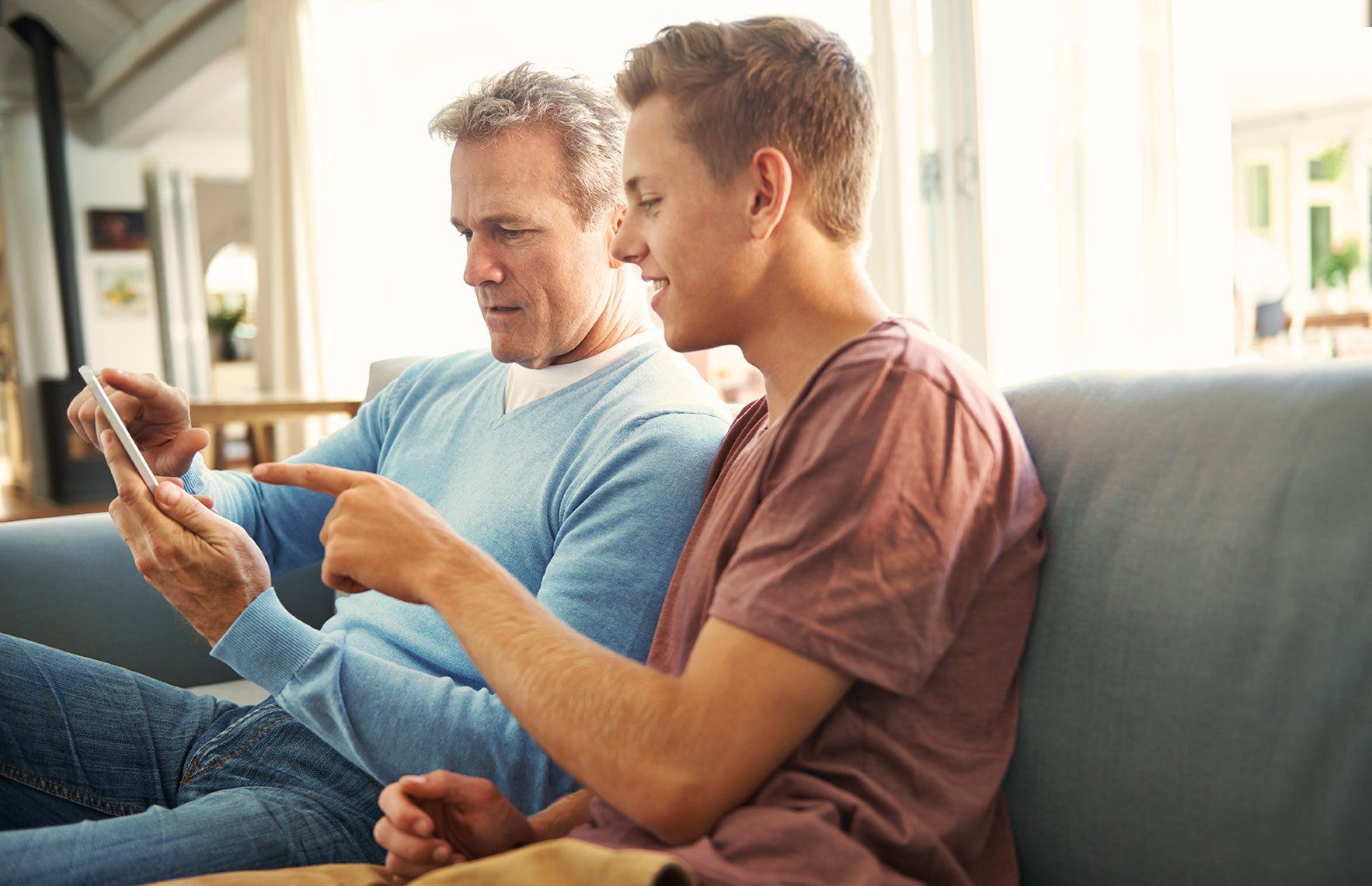Middle aged Caucasian dad wearing a light blue sweater and jeans showing his son, who is wearing a reddish brown shirt, his phone screen while sitting on the couch at home 