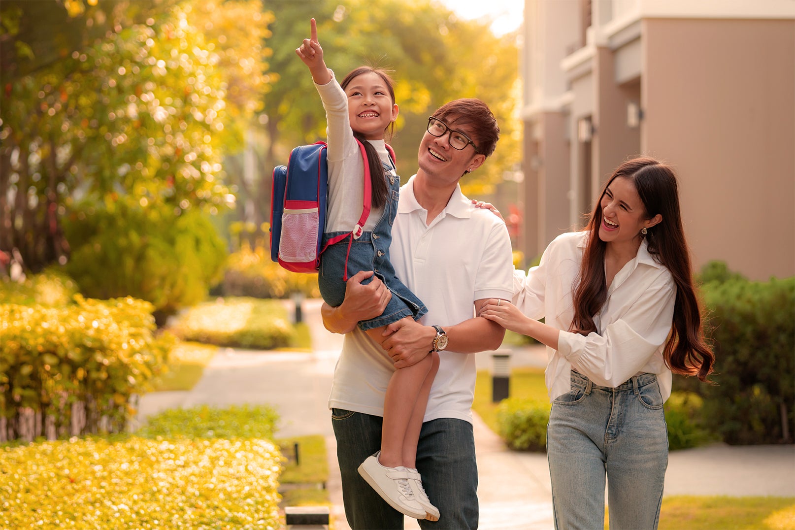 Young Asian man holding his daughter wearing her school backpack. He is walking with his wife in their neighborhood. 