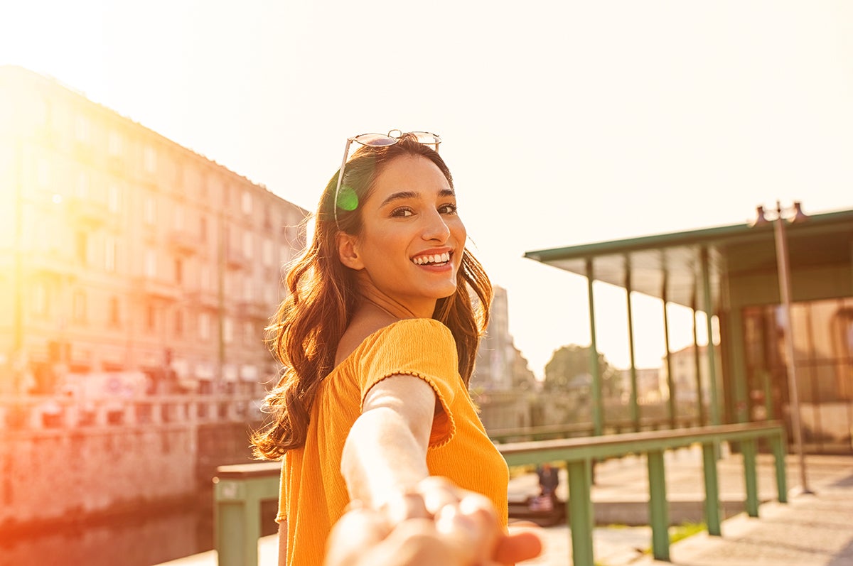 Young woman with brown hair wearing a yellow dress holding a friend's hand while walking into a foreign city 