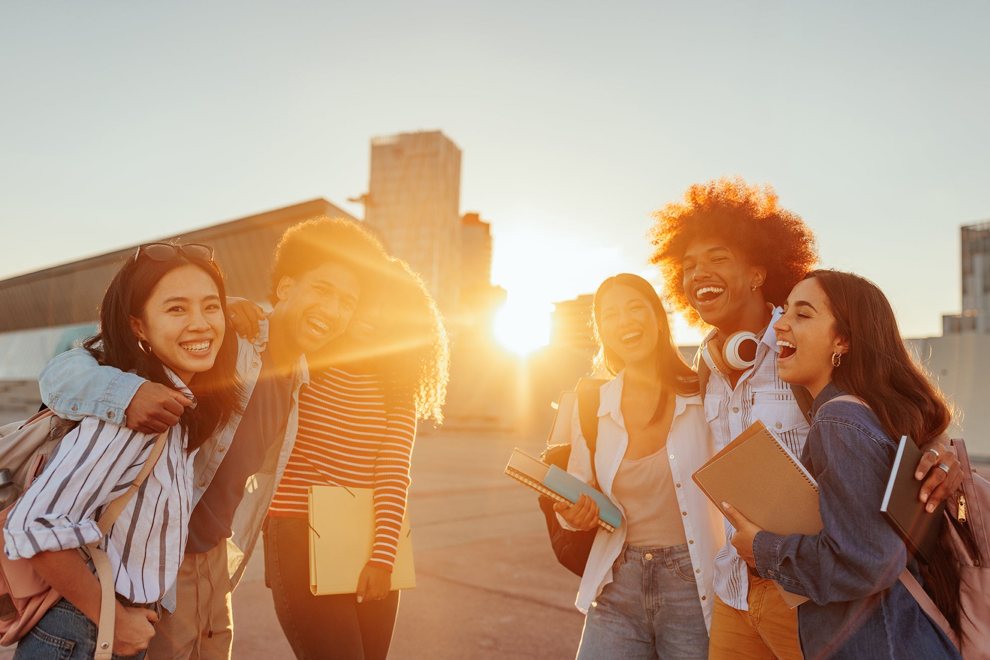 Group of five racially diverse young students holding notebooks and hugging as the sun sets on campus 